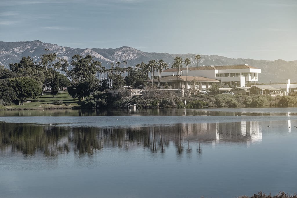 UCSB campus lake reflects modern buildings, palm trees, and mountains.